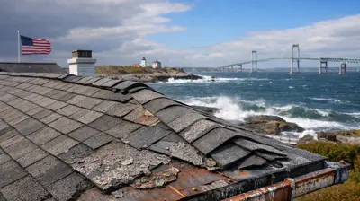 Techo desgastado por el clima de una casa costera en Jamestown, Rhode Island, que muestra la erosión de las tejas y los tapajuntas metálicos oxidados, con el puente de Newport y las olas del mar al fondo.